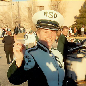 Woman smiling, standing outside on a sunny day, a shadow on the left, she wears a marching band uniform of white and green with MSU on the cap and an S on the left chest of the jacket.