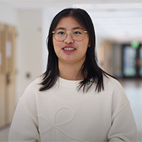 person with shoulder-length dark hair and glasses, wearing a white sweater, stands with a long hallway in the background.