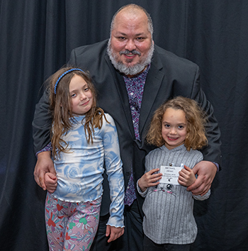 An adult person stands behind two young girls, leaning down toward them with his arms around them. All are smiling.