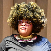 person with bushy black hair with blond highlights, wearing glasses, a black shirt, and large colorful necklace.