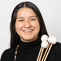 person with long dark hair and colorful necklace holds three drum mallets. 