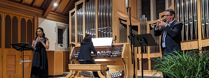 in a chapel with pipes of a pipe organ in the background, a person on left plays oboe, a person in center plays organ, and a person on right plays trumpet.
