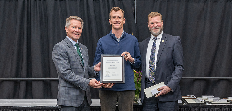 Three people wearing business suits smile while the person in the center holds a framed certificate