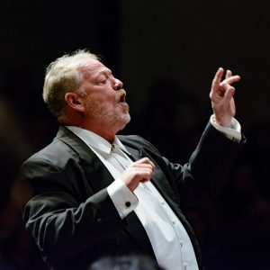 person in tuxedo raises arms while conducting music.