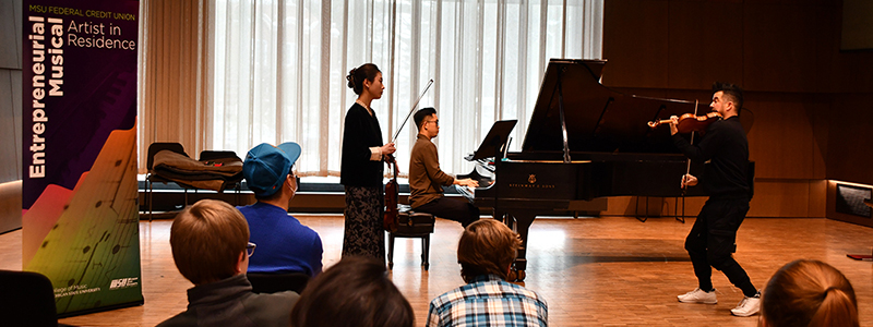 people in a large music rehearsal space, some watching while another at a piano and a person holding a violin watch another person demonstrating something on a violin. A banner on the left reads "MSU Federal Credit Union Entrepreneurial Musical Artist in Residence"