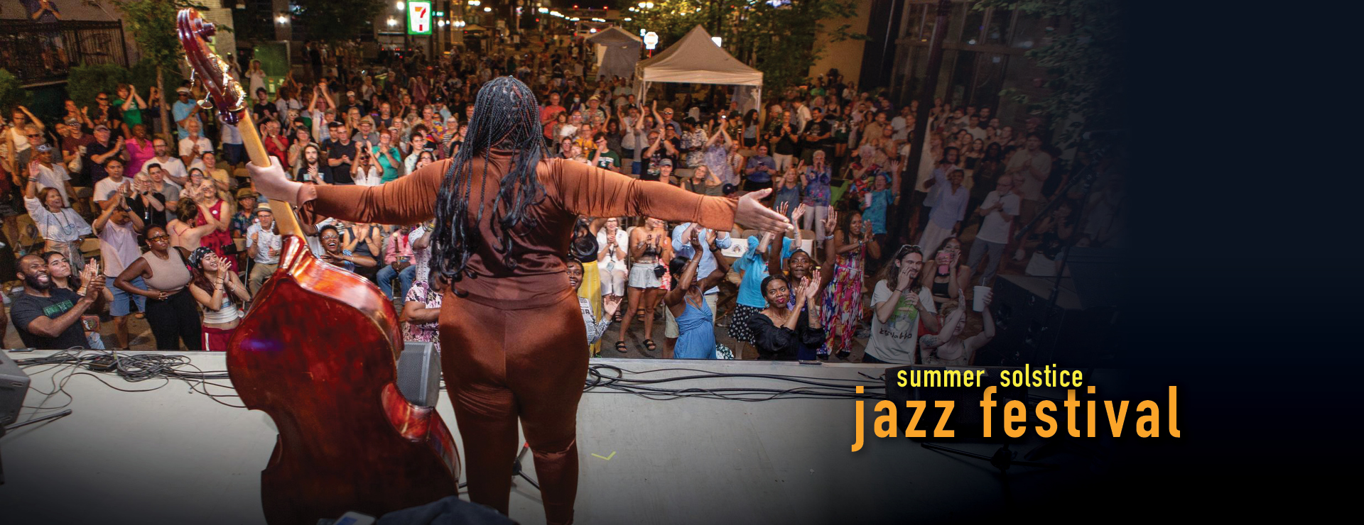 Summer Soltice Jazz Fest: Image of jazz bassist on stage, arms extened outward with a full crowd in the street.