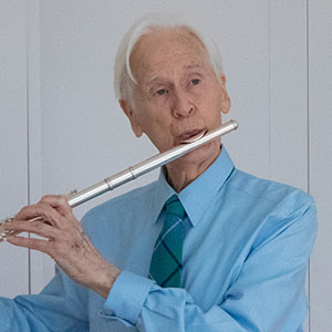 person with white hair wearing light blue shirt and green tie plays a flute