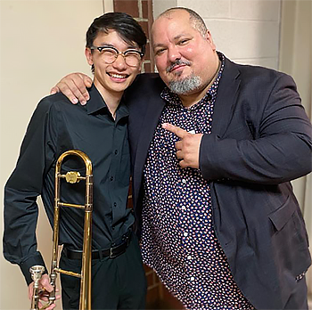 a younger person on left holds a trombone while the older person smiles and points at the younger person.