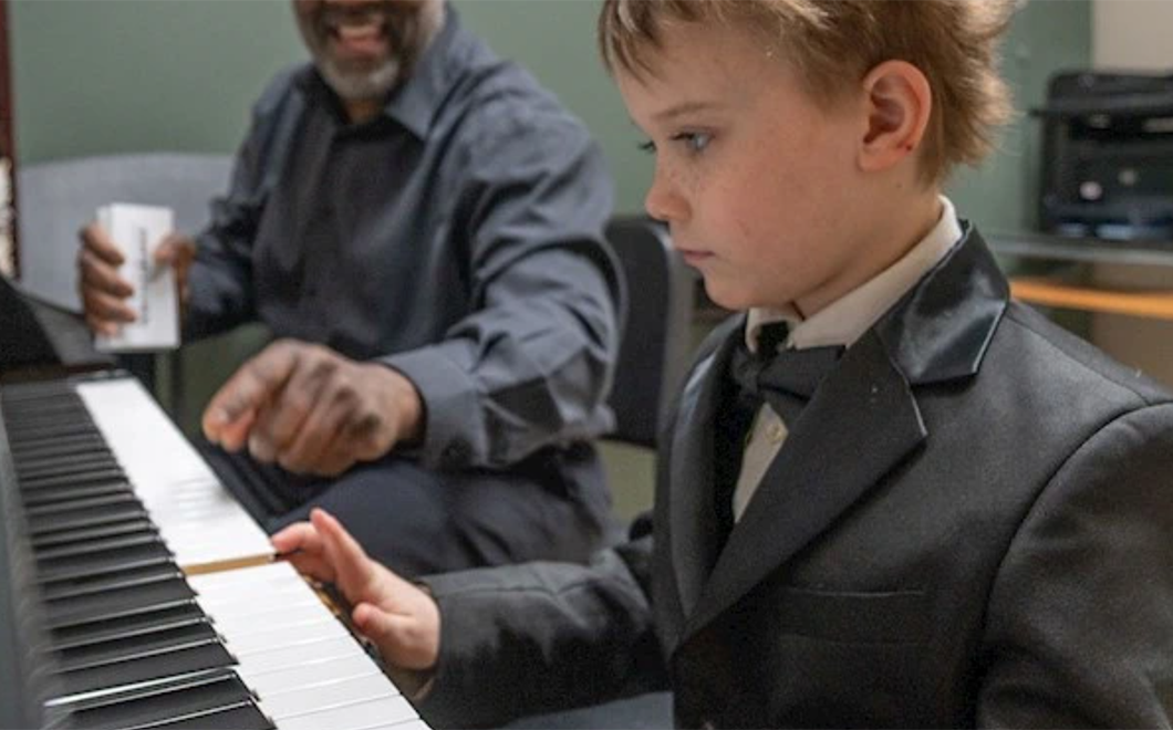 A young child wearing a suit with a bowtie plays the piano while an adult in the background smiles and points to the keyboard.