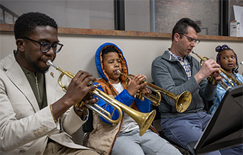 Two adults and two children sit on chairs in a room, each playing a trumpet.