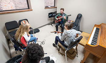 An instructor sits in a room with four students, each holding a guitar. A piano is on the right.
