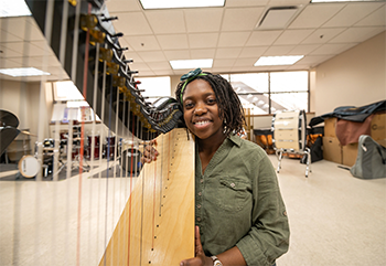 In an expansive, well-lit room with music-related items in the background, a young person smiles while sitting with a harp.