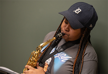 seated person plays saxophone, olive green wall behind. The person wears a dark blue baseball cap with the Old English D of the Detroit Tigers.