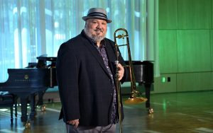 person standing in large performance hall wearing a suit jacket and hat while holding a trombone; a green hue, curtain, and piano in the background.