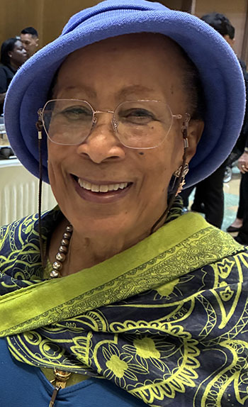 a smiling person with glasses wearing a lavender hat with green clothing