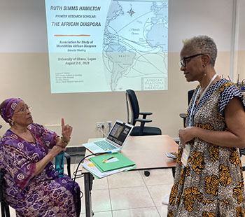 a seated person on the left speaks to a person standing on the right with a projected image of a presentation on the wall behind them