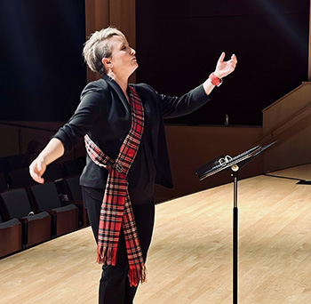 person on stage wearing all black with a red plaid sash, arms out and raised while conducting