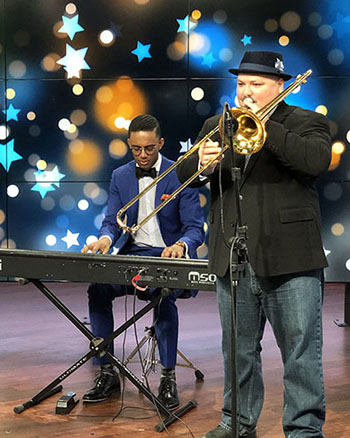 seated person in suite and bow tie plays an electric keyboard while a person standing, wearing a suit jacket and hat, plays trombone