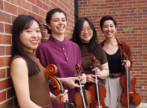 Red Cedar quartet members, smiling and looking at camera, standing against decorative brick wall. Cellist, two vionlinists and a violist.