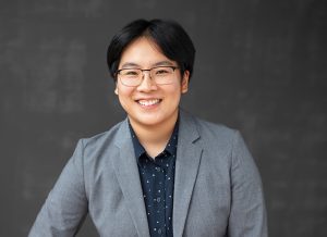 smiling person in glasses and a gray suit coat with dark shirt