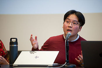 Person in red sweater and glasses speaks into a microphone