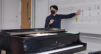 person standing and pointing to musical notes on a white board with a grand piano in the foreground