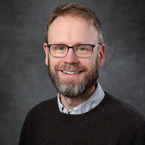 person with beard wearing glasses and brown sweater with white collared shirt underneath, smiling