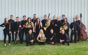 Chamber wind ensemble group of more than a dozen instrumentalists, photographed outside standing against a pole-barn building on grass