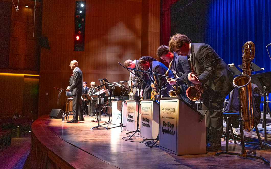Musicians of Jazz Orchestra 1 take a bow during a performance at Fairchild Theatre. Blue colored curtain in background with ambient lighting on stage. Conductor standing at the front