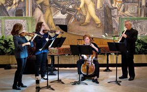 Members of L’Invenzione perform in the Rivera Court of the Detroit Institute of Arts. Five musicians playing string, flute and harpsichord instruments, artistic murals are in the background.