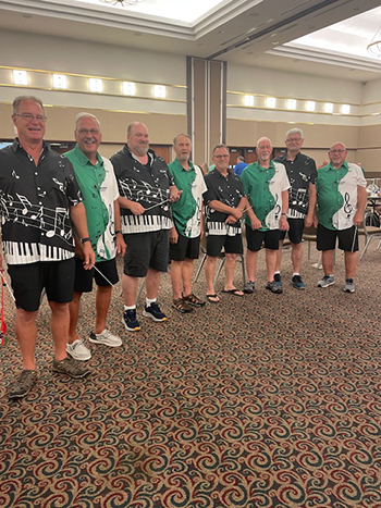 eight people stand in a row, each smiling and wearing different shirts with musical symbols on them