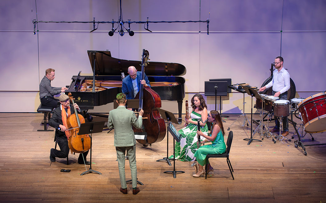 American Modern Ensemble musicians performing on stage, dramatic lighting with conductor directing a pianist, cellist, bassist, bassoonist, clarinetist and percussionist.