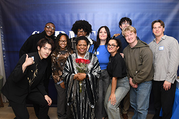 a person holding roses and smiling stands in front of a blue curtain surrounded by young adults also smiling