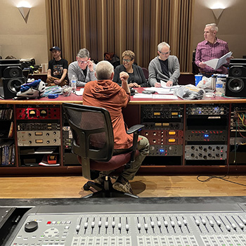 In a recording studio, a mixing board in the foreground and peolpe around a table in the background