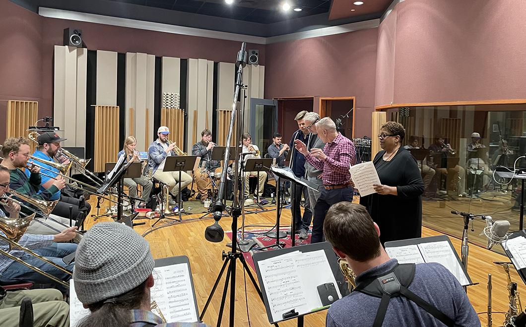 a group of musicians in a recording studio with a long row of seated horn players, a standing person who is singing, and tow other people following along with sheet music