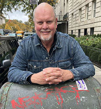 person with white goatee outside wearing a denim shirt and leaning on a graffitied mailbox