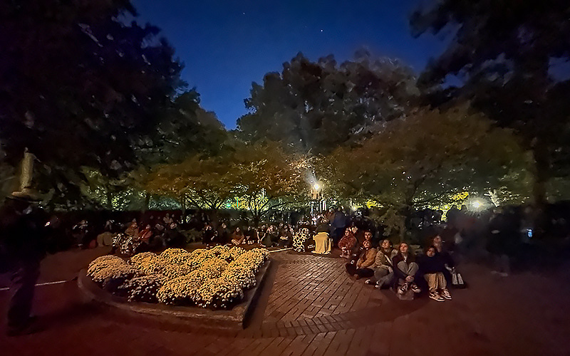 a group of people sit outside looking in the same direction while a bright projector light shines behind them