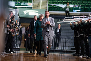 a single file line of three adult people walking into a gymnasium with band musicians playing on either side of them.