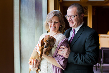 a couple stands close together while one person holds a small dog that they are both smiling at