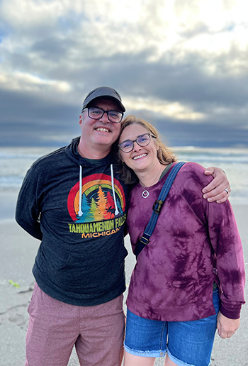 two people smile with arms around each other while standing on a beach with water and clouds in the background