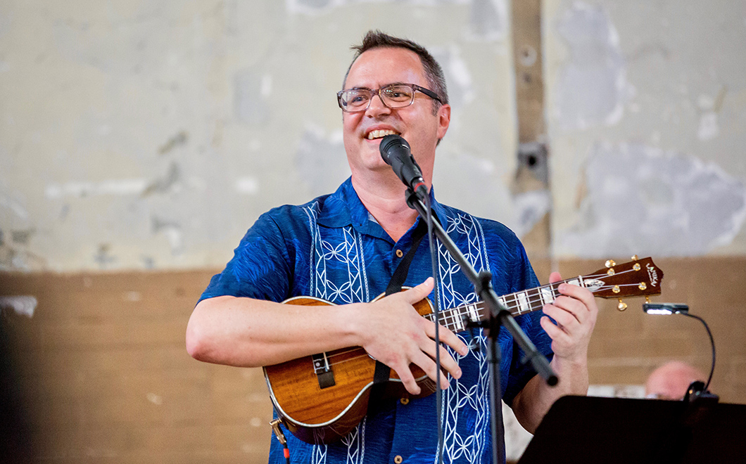 person in blue shirt in front of a microphone smiles while playing ukelele