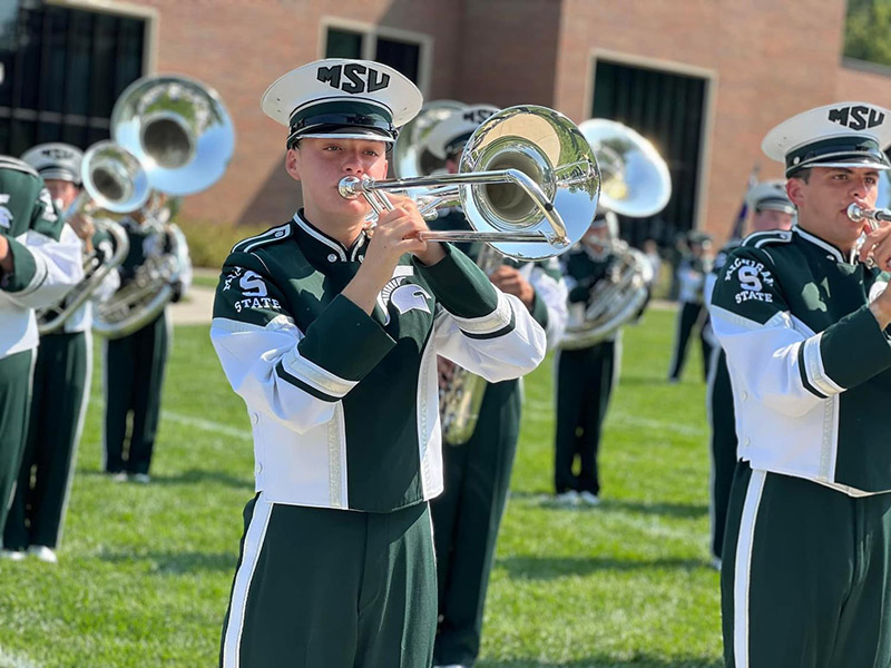 people in green and white marching band uniforms stand on a field while playing trombones