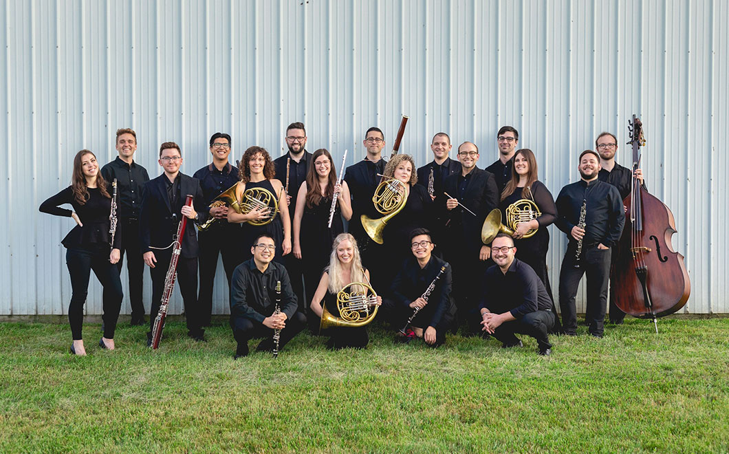 the Newfound Chamber Winds poses on grass outside of a polebarn. Each poses with their instrument, about 20 musicians