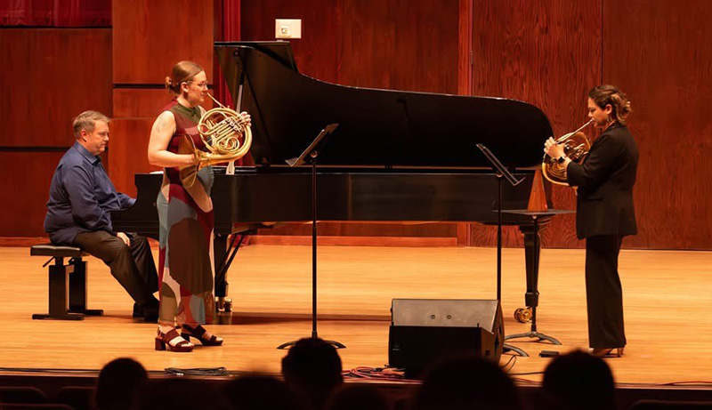two horn players perform on a stage with a pianist