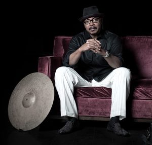 Stylized portrait, jazz drummer Harvey Mason. Seated at burgandy colored couch, clasping hands, reflective expression on face. drum symbol leaning against couch