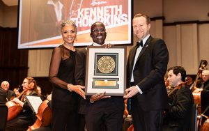 three people on a stage, the one in the center holds a framed award