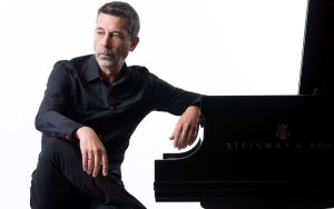 Portrait: Pianist Sergei Kvitko, casually seated at piano, turned towards camera with elbow leaning on piano, thougthful facial expression looking away from camera