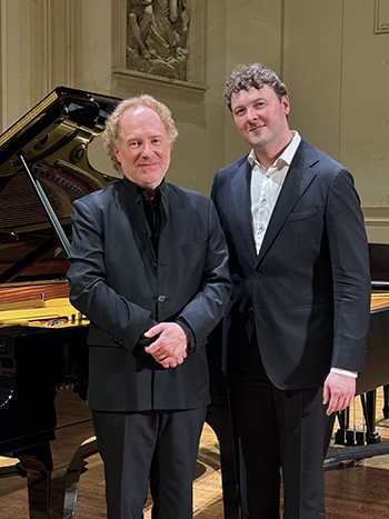 two people in suits stand in front of a grand piano