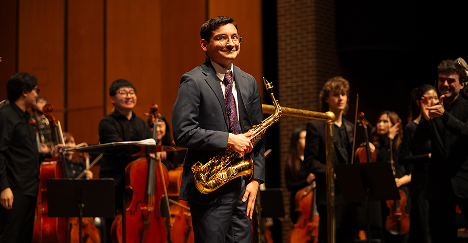 person with saxophone smiles on stage while other musicians are in background