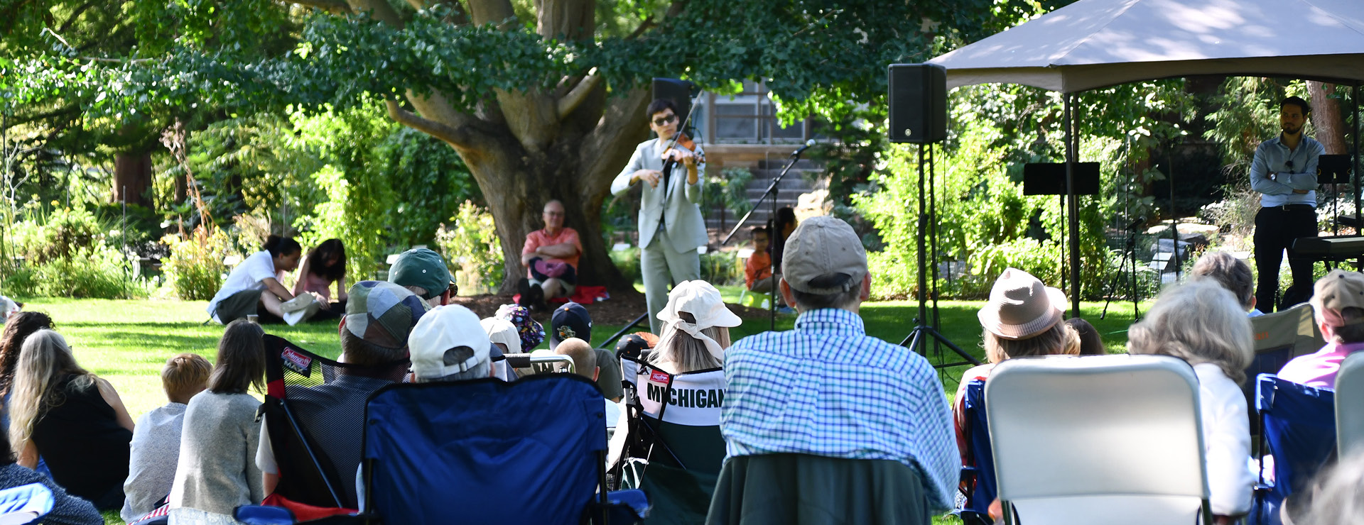 Audience members sit on folding chairs and blankets on a lawn while a musician performs outdoors beneath large trees in a garden setting.